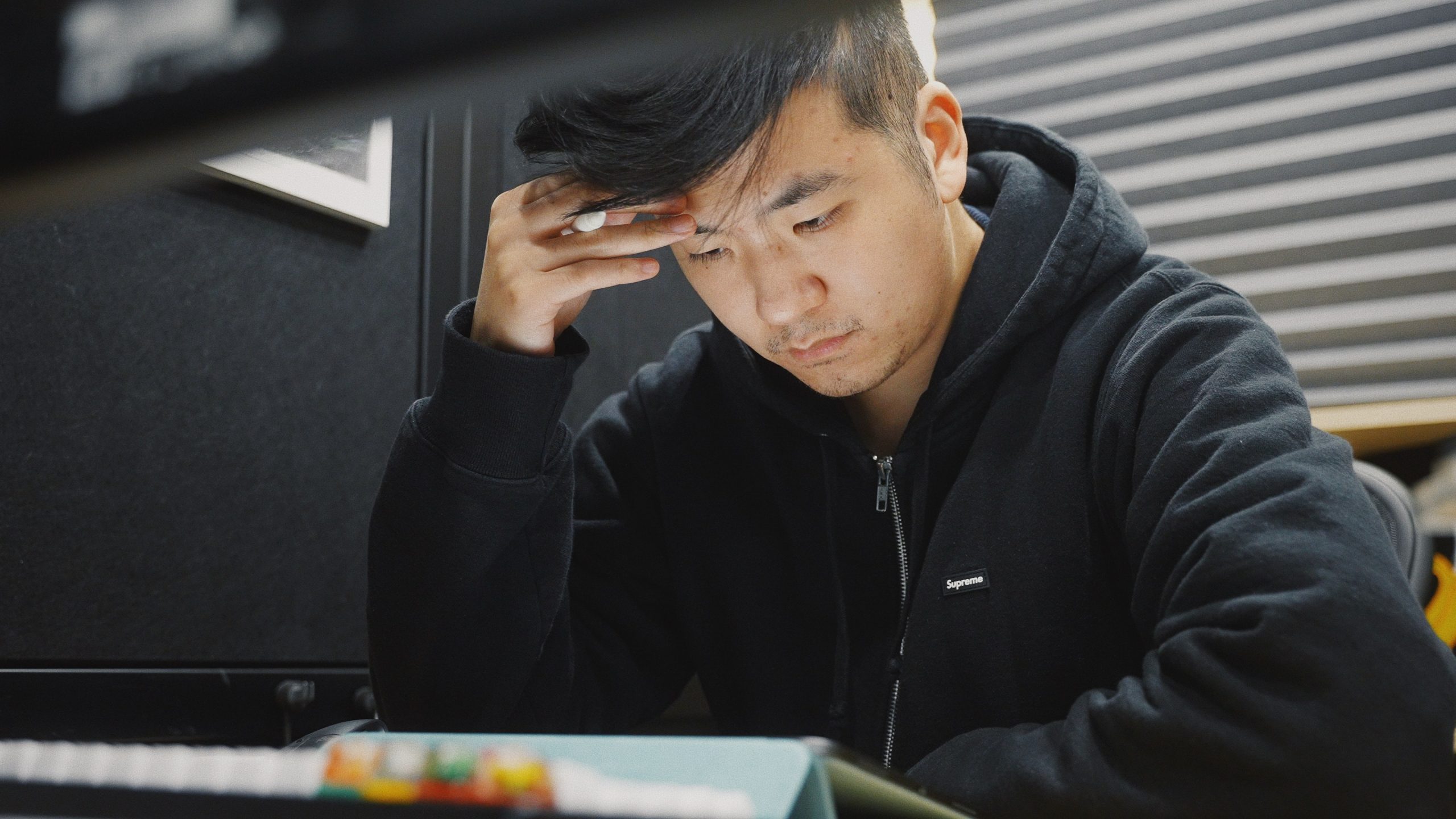A young Asian man wearing a black hoodie looks thoughtful as he sits on a desk.