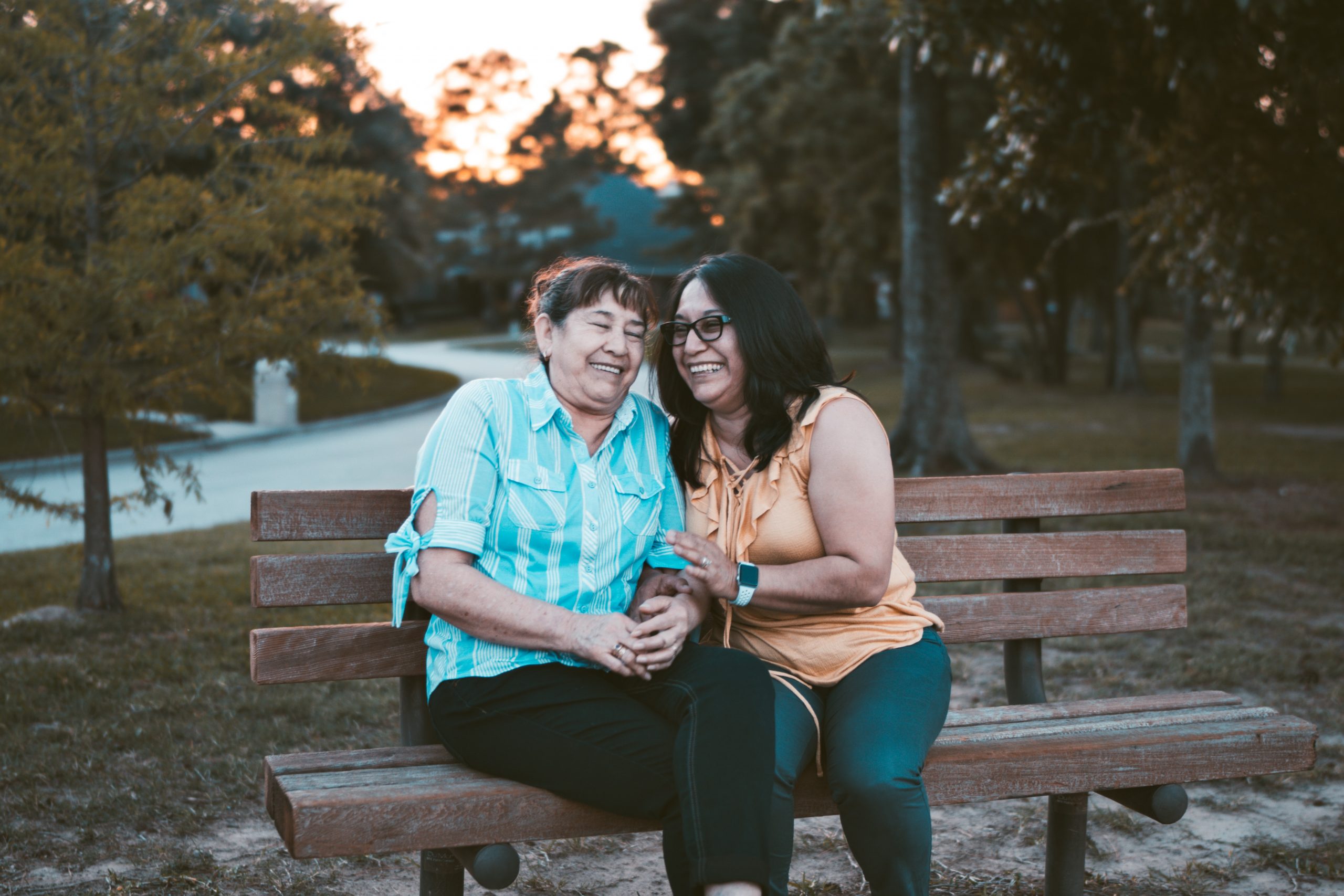 Two middle aged Hispanic women sit on a bench and laugh together