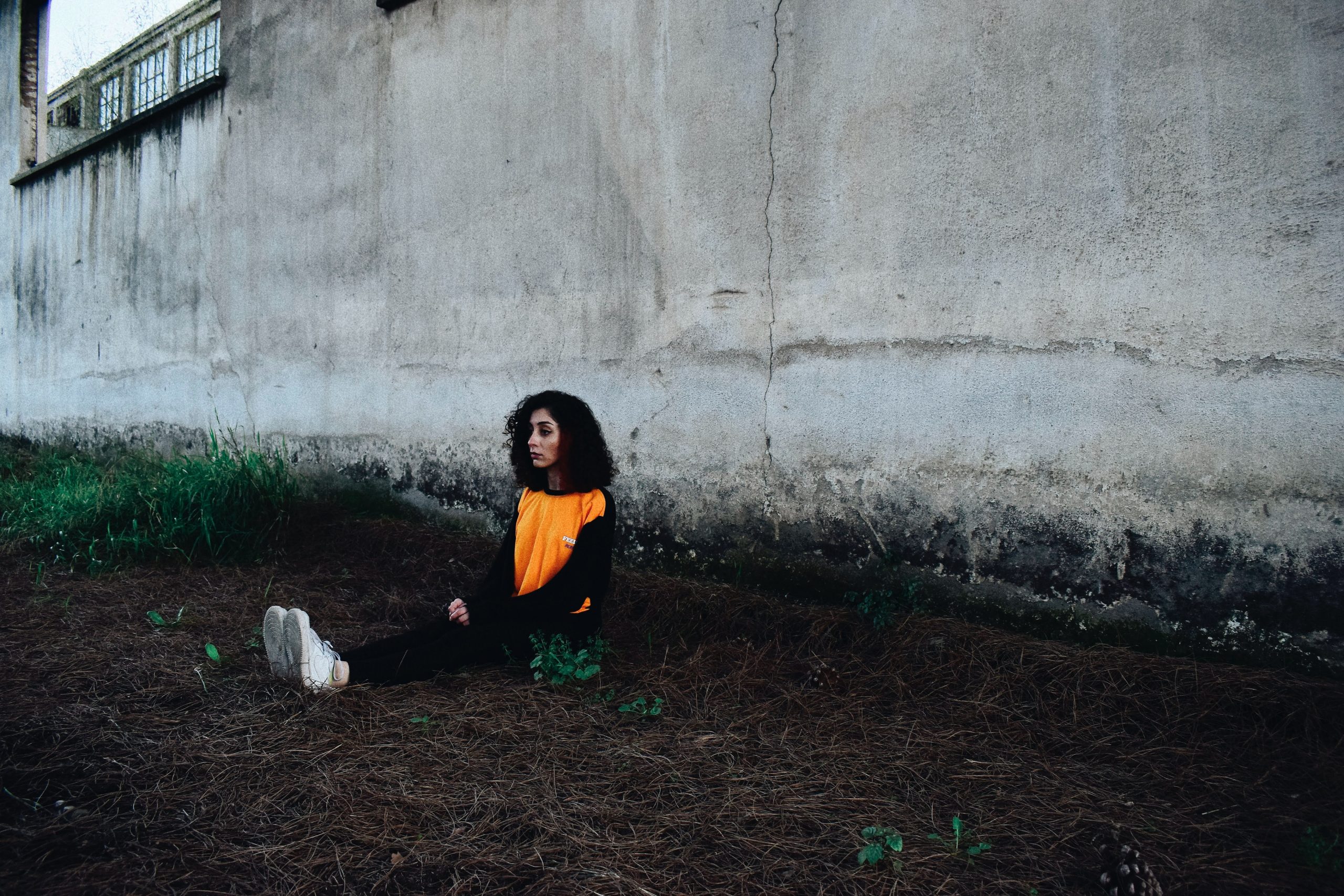A young mixed race girls sits in th dirt under a concrete wall. She looks sad and wears an organge t-shirt and dark trousers