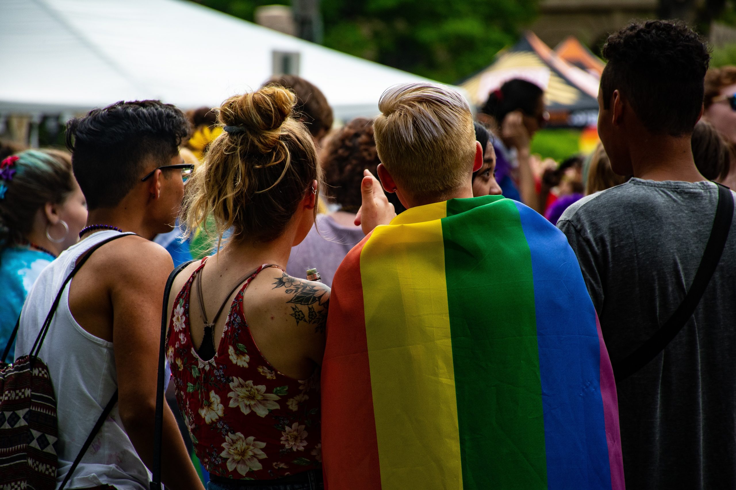 The photo focuses on the backs of a group of friends at a Pride event. One is draped in a Pride flag