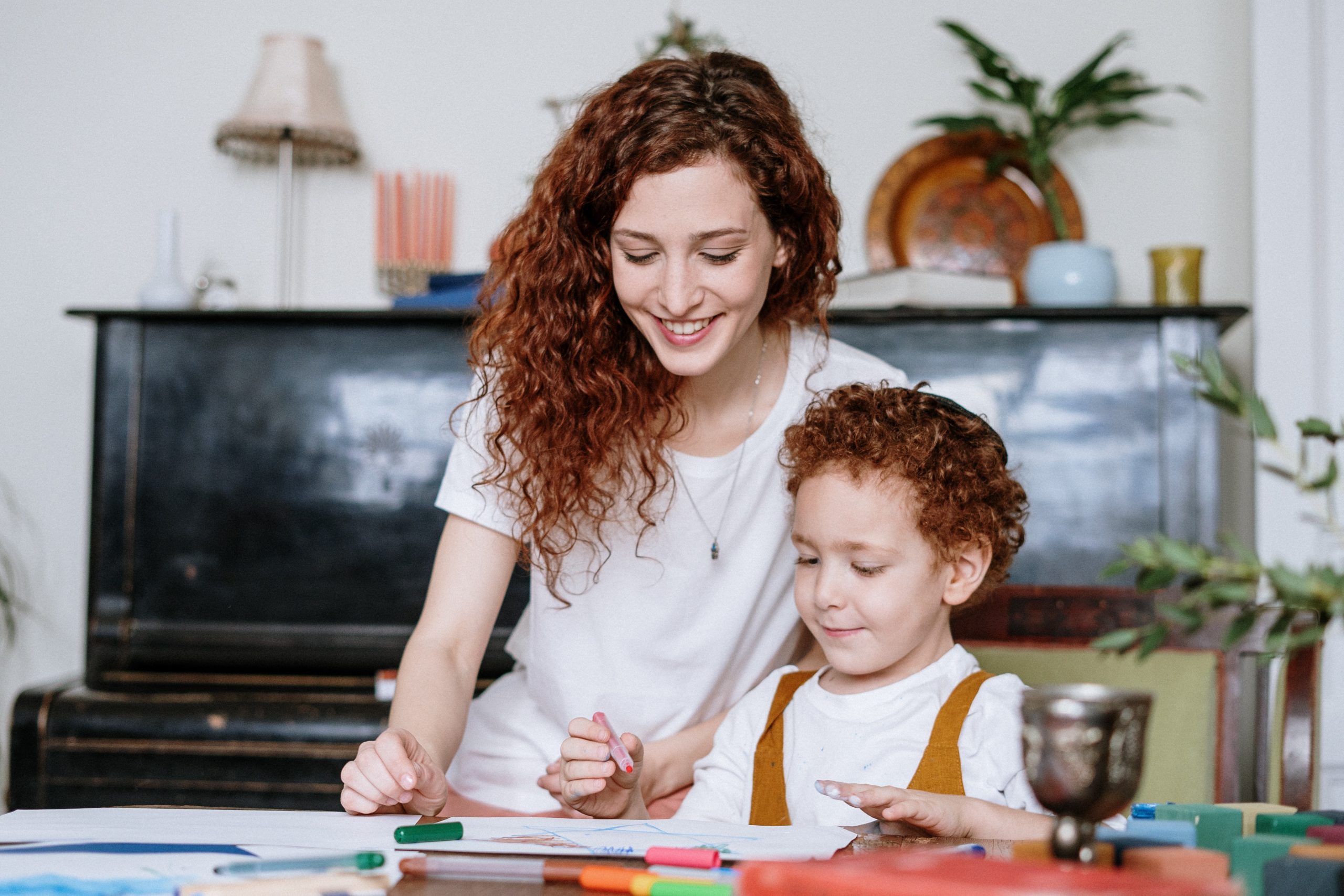 A young mum with curly red hair sits and plays at a able with her young son, who has matching red curly hair. 