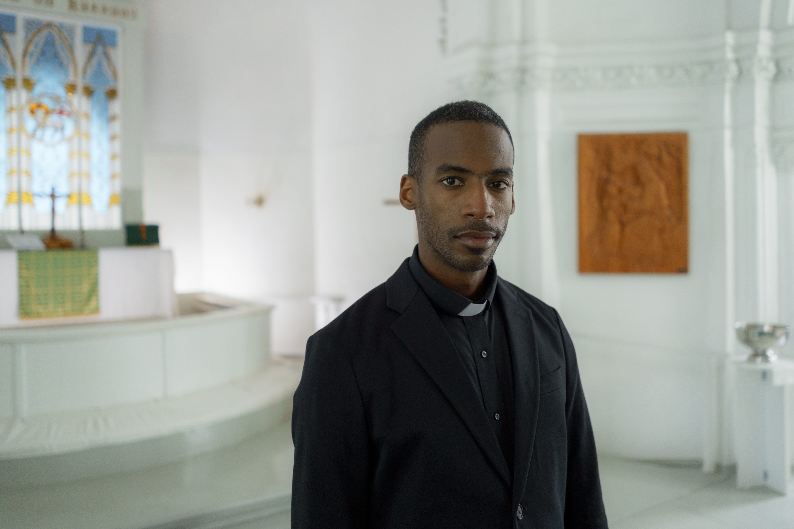 A Black priest wearing a black top and white collar looks directly into the camera in his church.