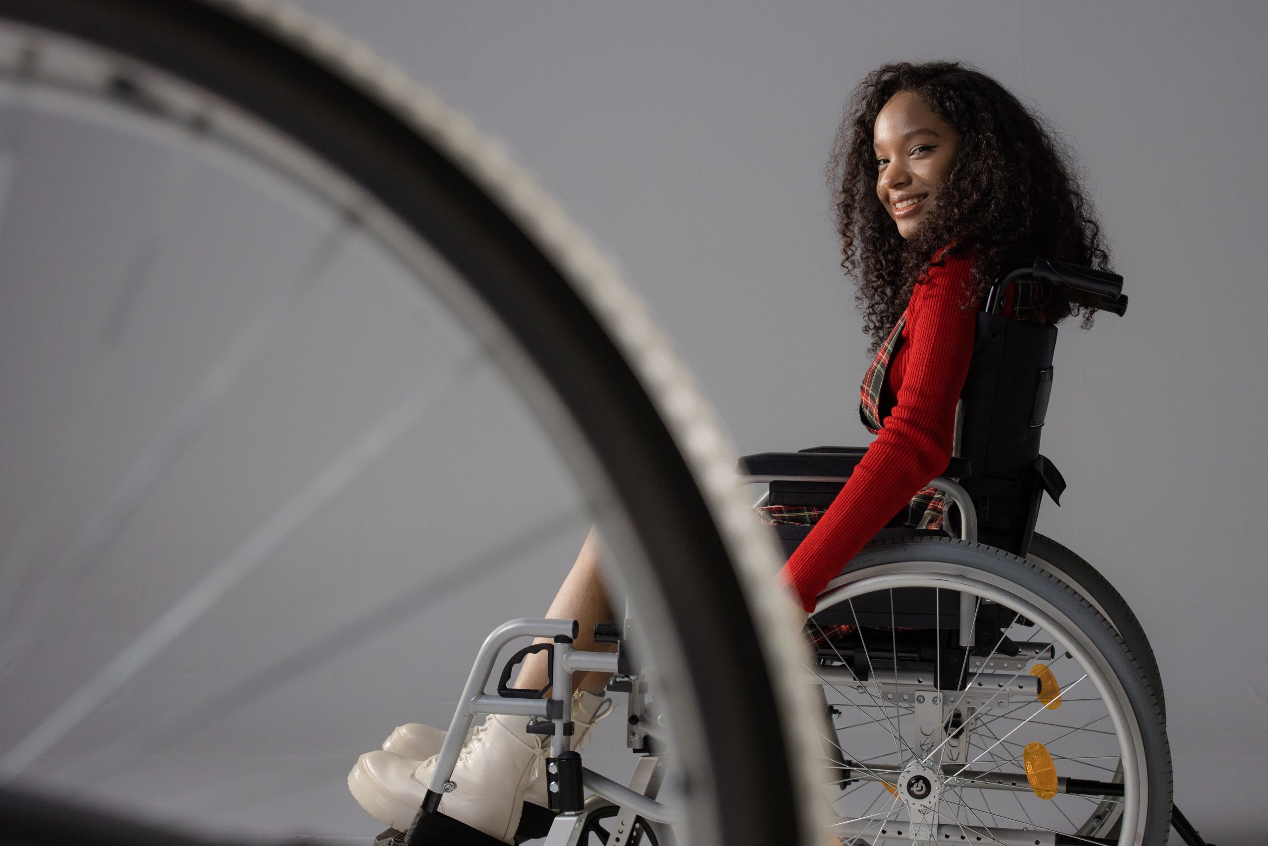 A young Black girl uses a wheelchair against a grey backdrop. She is smiling and wearing a red long sleeved top