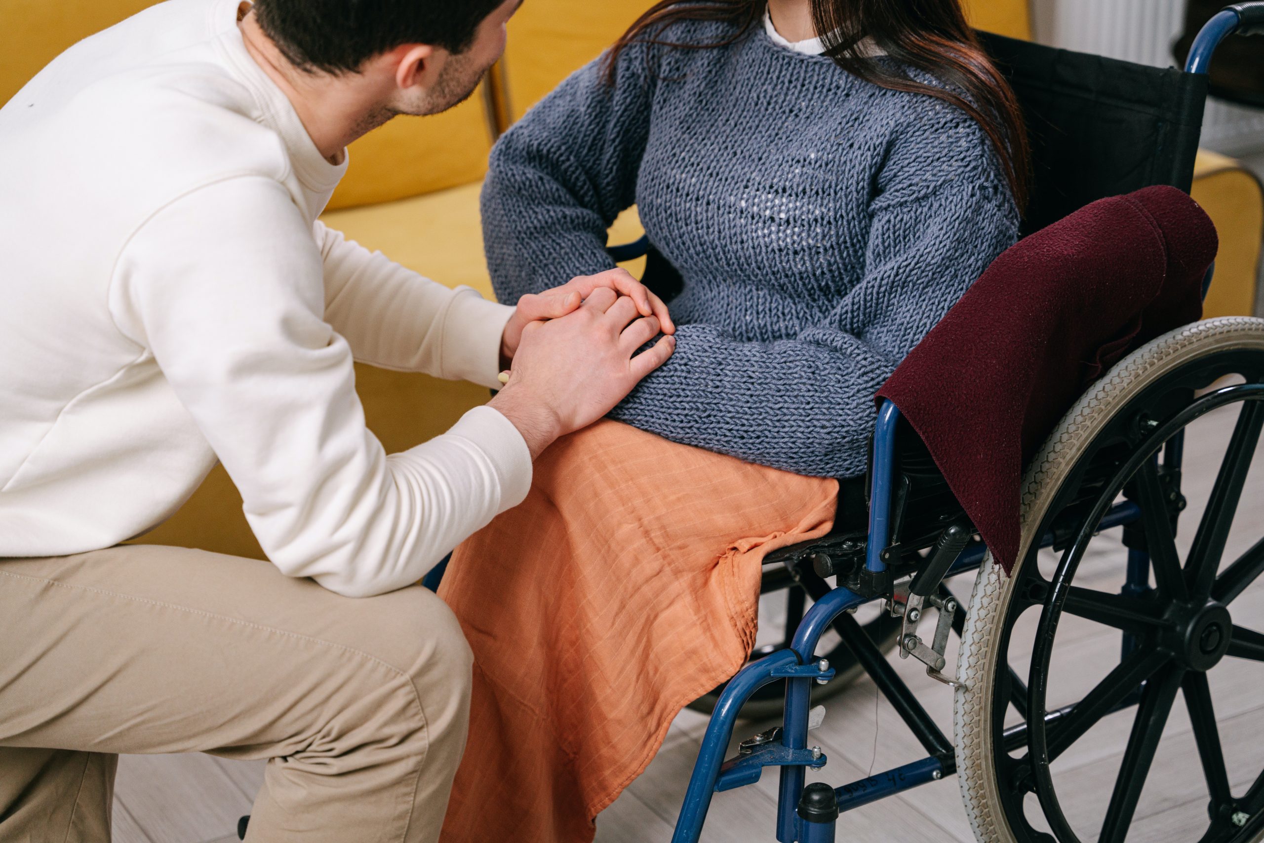A person in a white shirt and tan trousers holds he hand of a wheelchair using woman