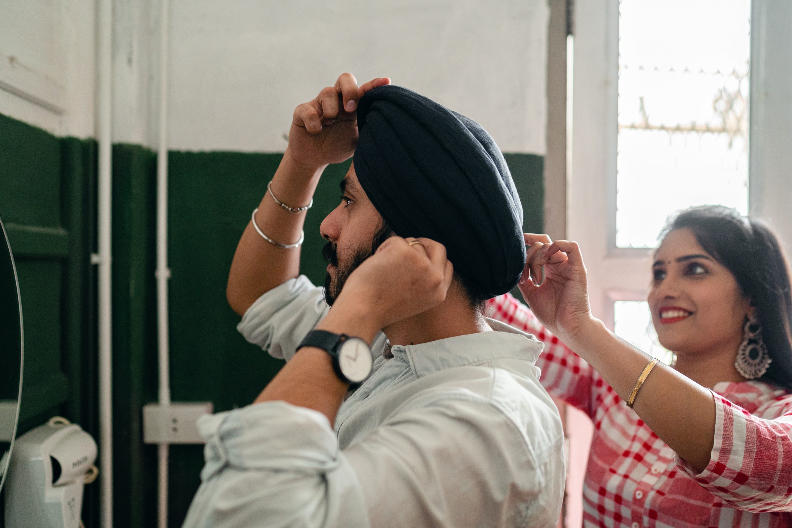 A woman helps her Sikh husband adjust his black turban in the mirror