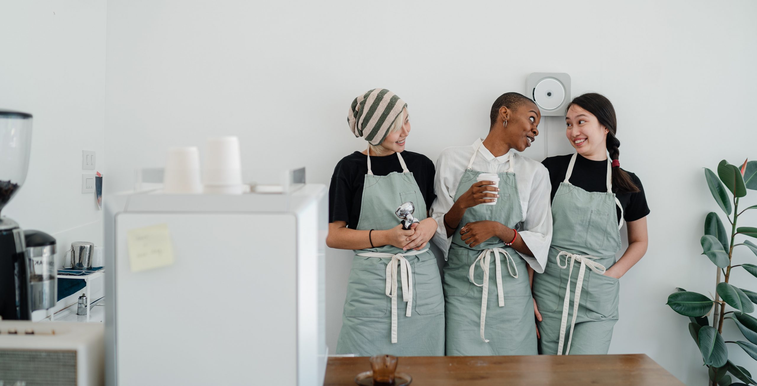 A group of three women wearing light green aprons lean against a wall and chat. One is white, one is Black and one is Asian. 