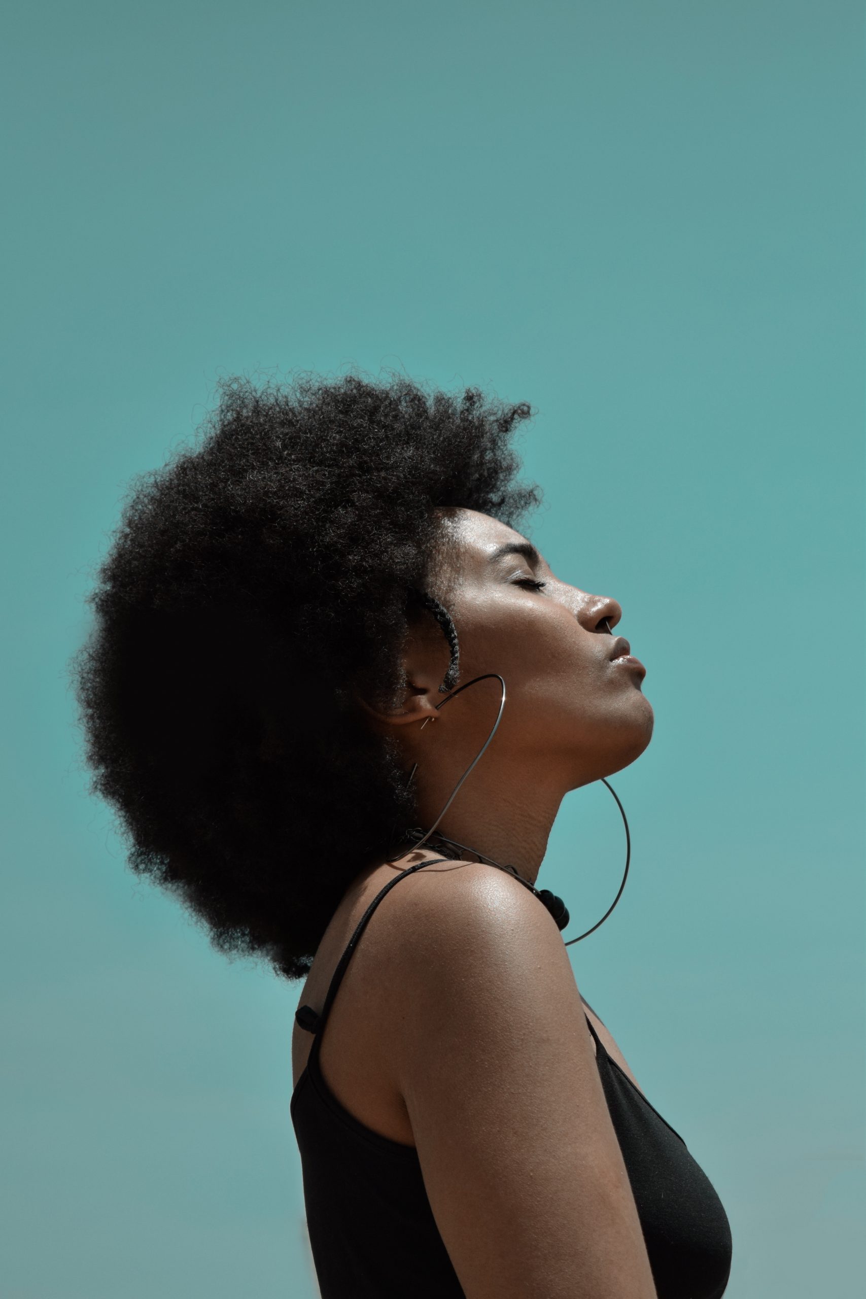 A Black woman with afro hair looks towards the sun with her eyes closed. It is a profile shot from the side showing her arm, shoulder and head.