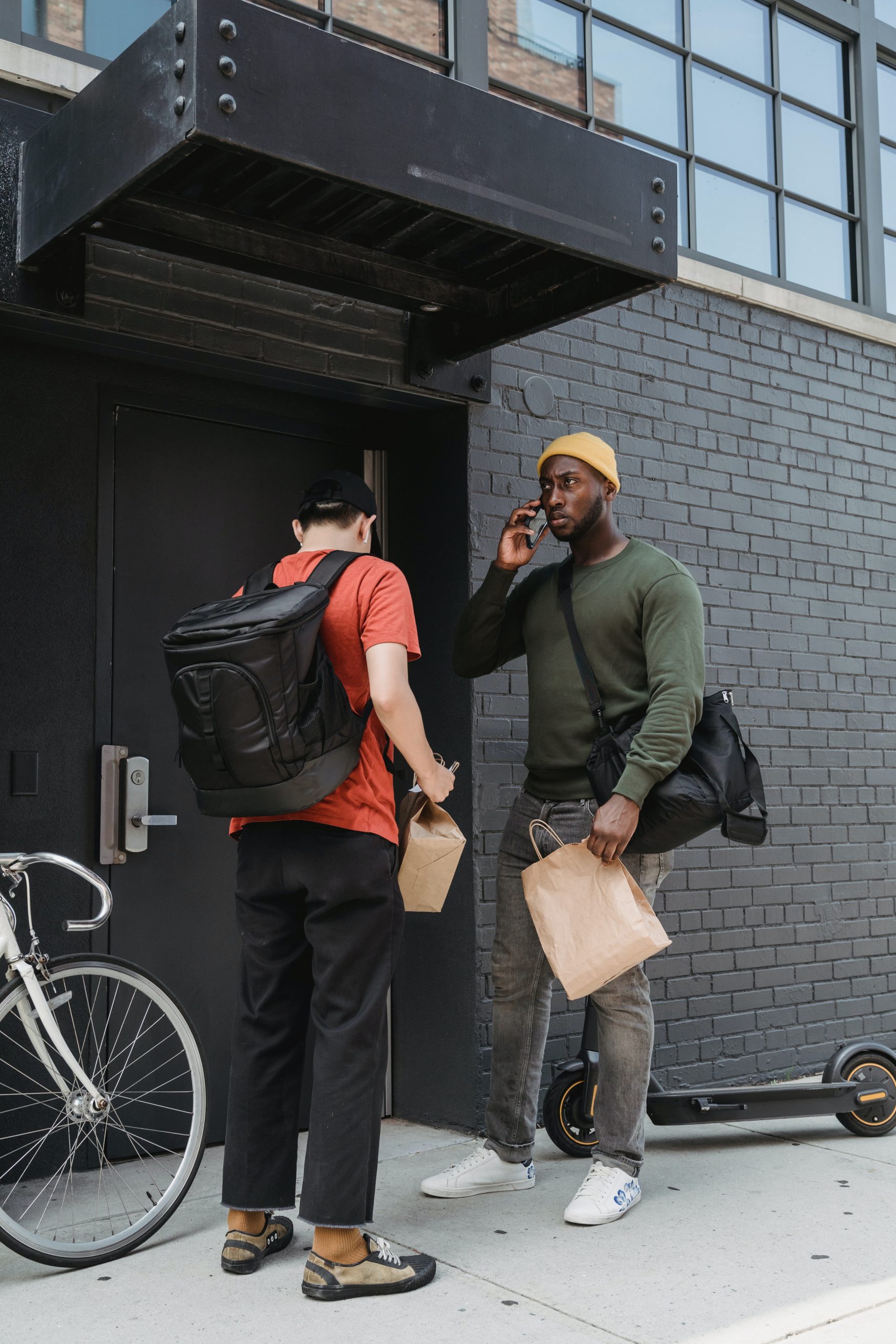 Two food couriers stand outside the entrance to an apartment building. One is on the phone while holding a brown paper delivery bag and the other is carrying a black backpack