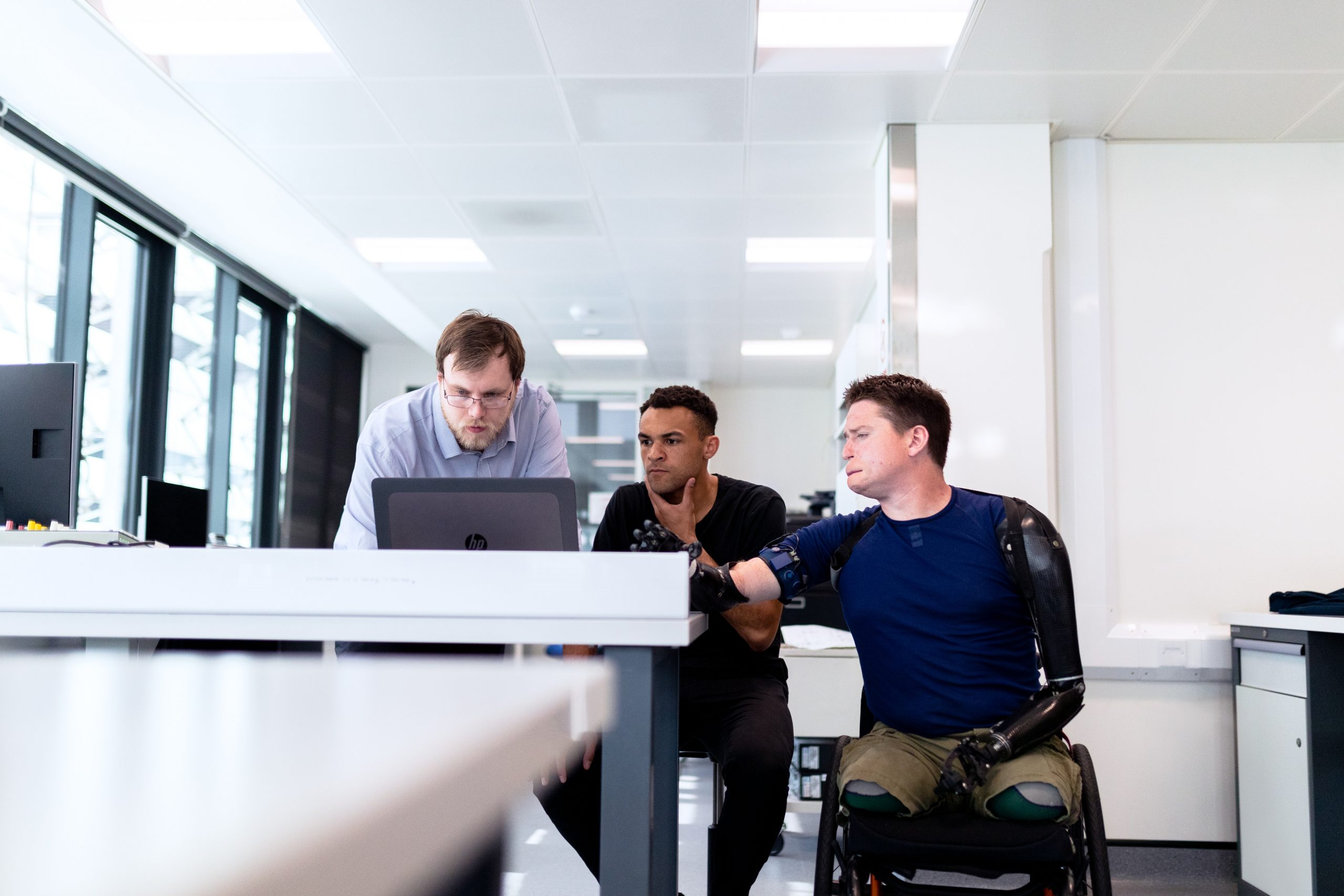 Three young men all gather around a desktop computer to work. The one on the far right is a multiple amputee and a wheelchair user.