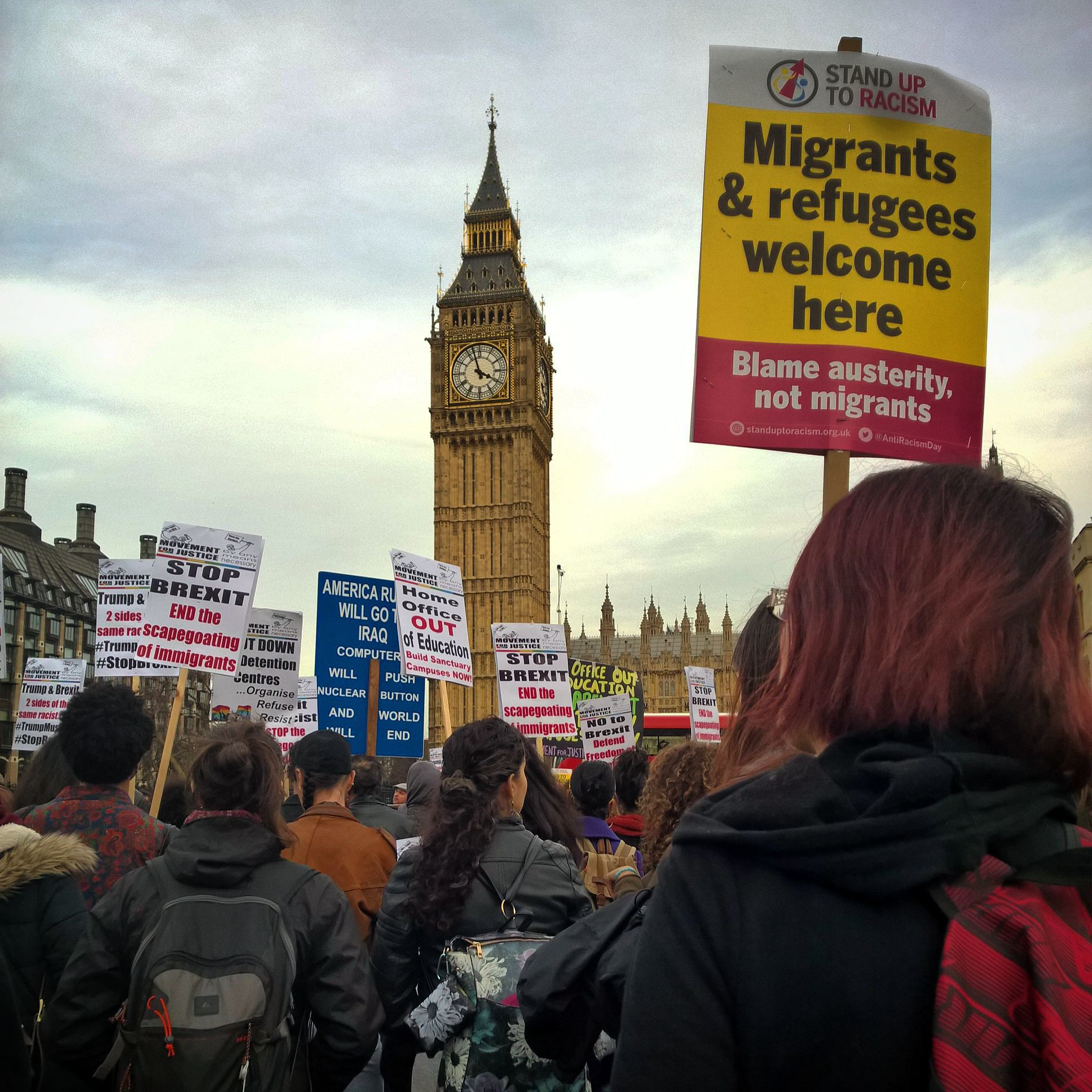 A protest sign reads "refugees and migrants welcome here" in black lettering on a yellow protest sign. The sign is held a loft a crowd of other protestors with the Big Ben clock tower looming in the background. 