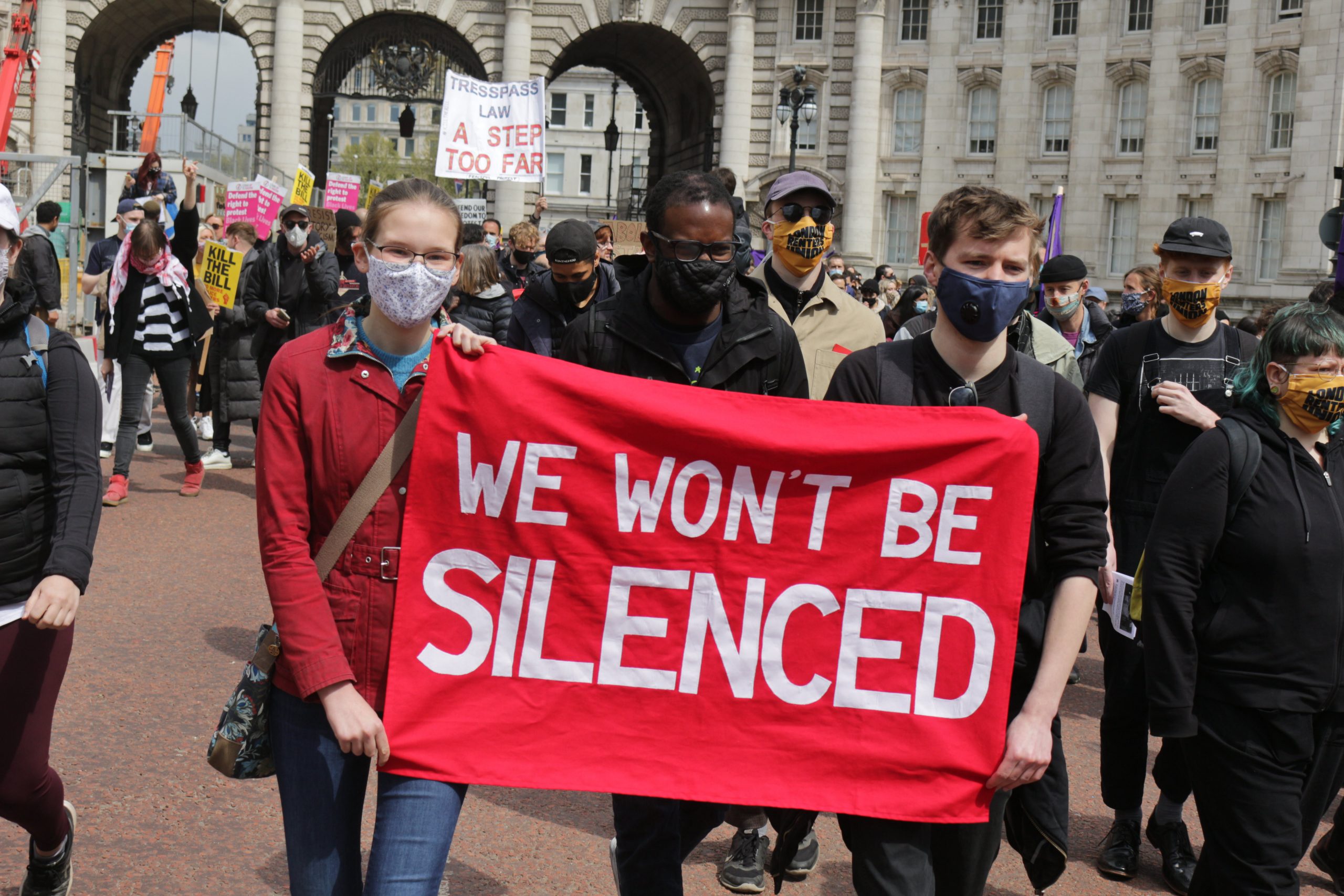 Three young protestors march through London on a Kill The Bill protest carrying a red banner with white writing on it which says 'We won't be silenced'