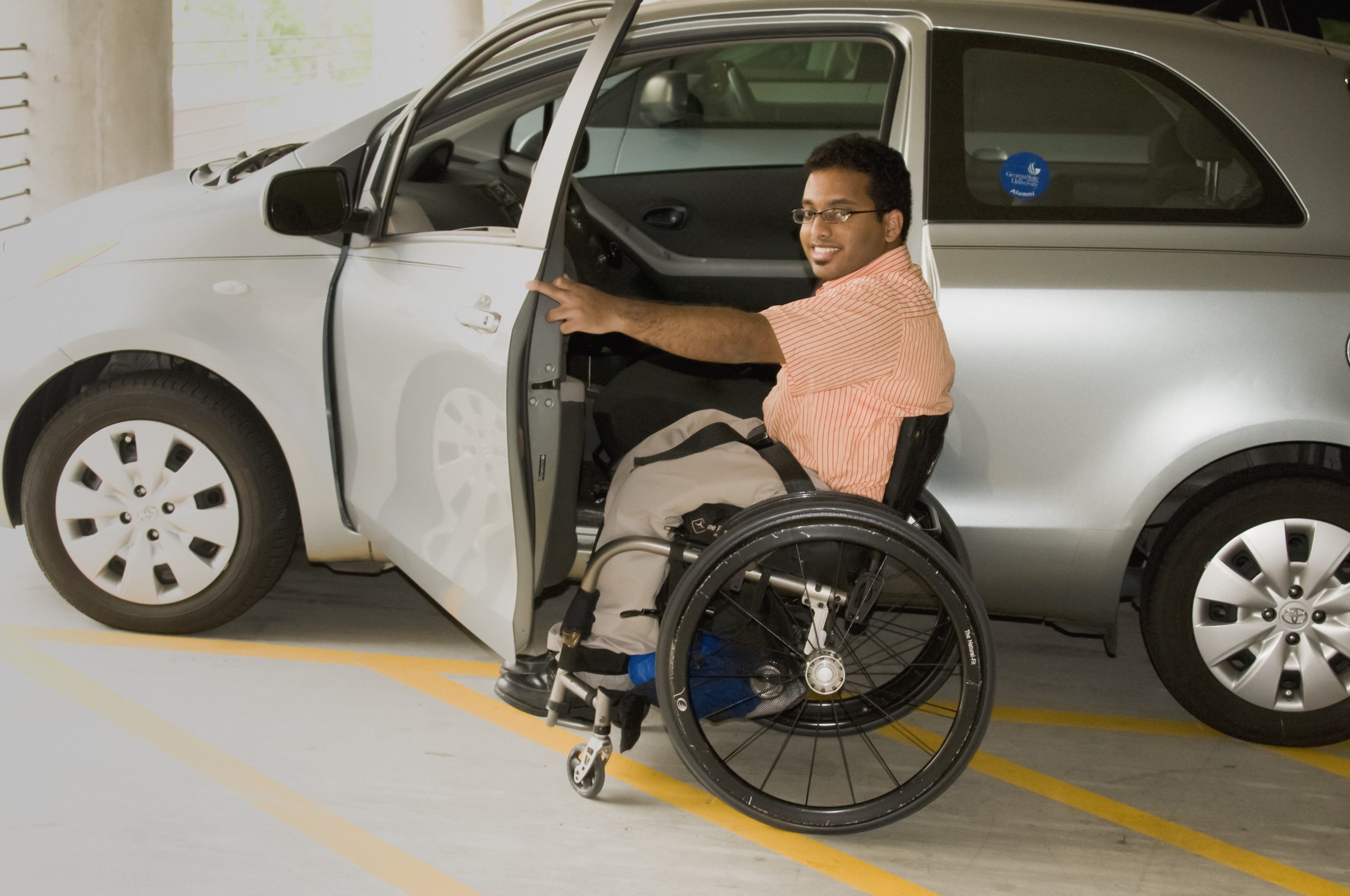A wheelchair user opens a silver car door and prepares to get in. He is wearing a peach shirt and light coloured trousers