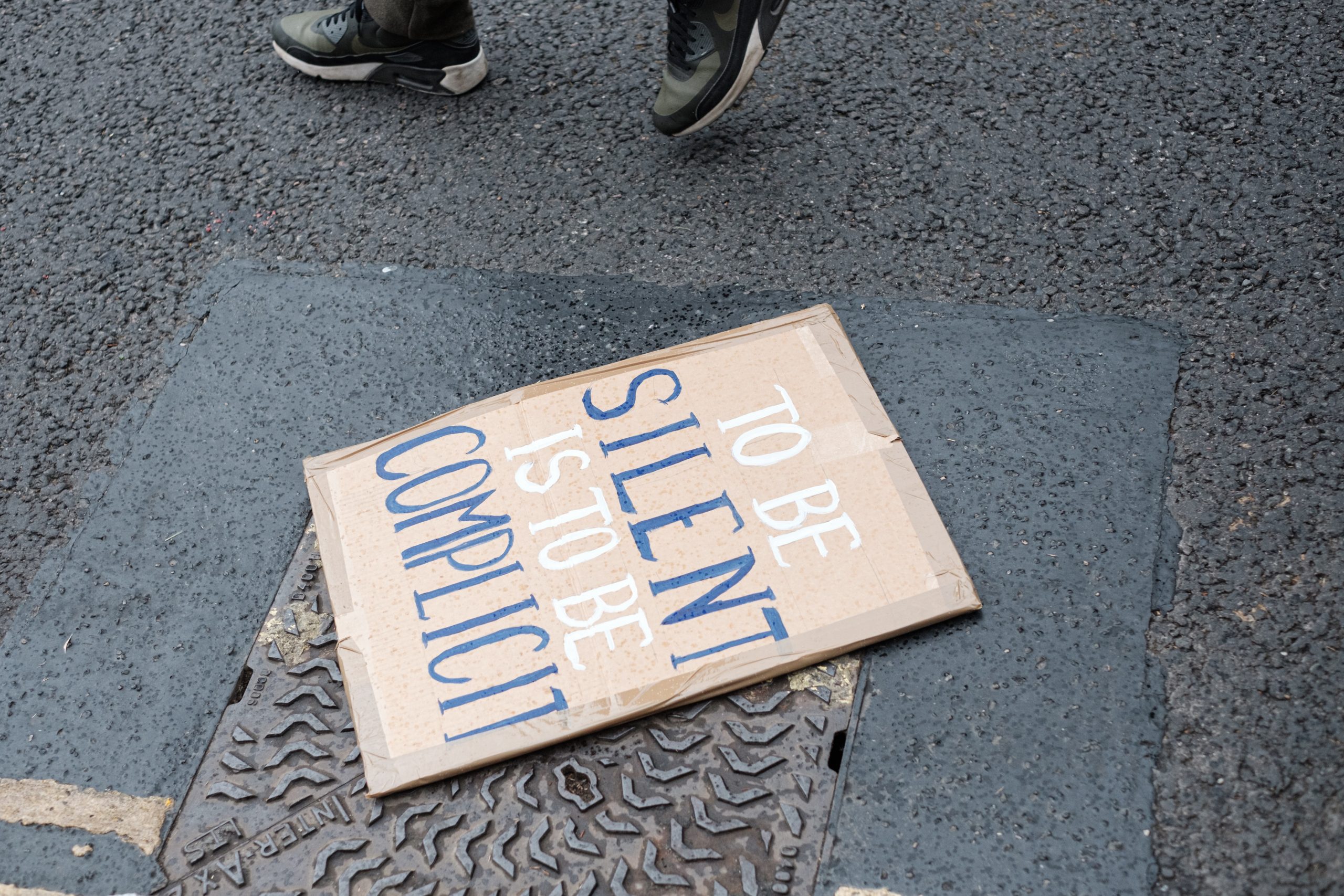 A sign written on cardboard lies on the floor. It reads "To be silent is to be complicit" in alternating white and blue text