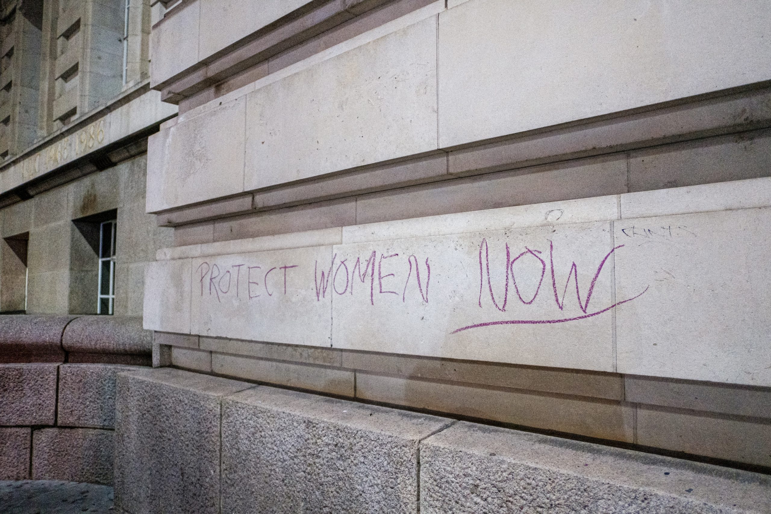 The words "protect women now" are written on a stone wall