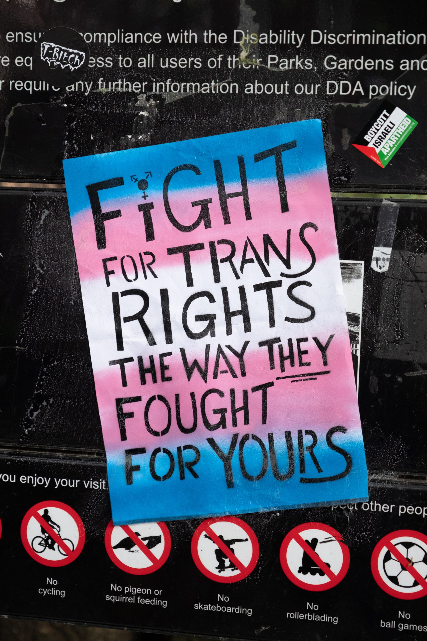 A protest sign painted in the trans flag colours reads "fight for trans rights the way they fought for yours" in black lettering.