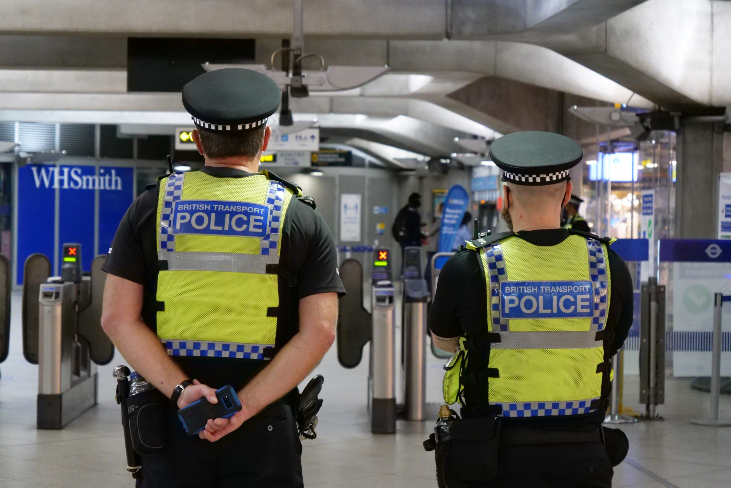 Two male police officers stand facing away from the camera in a London Underground station. They are both wearing yellow high vis jackets with the word "police" emblazoned on the back.