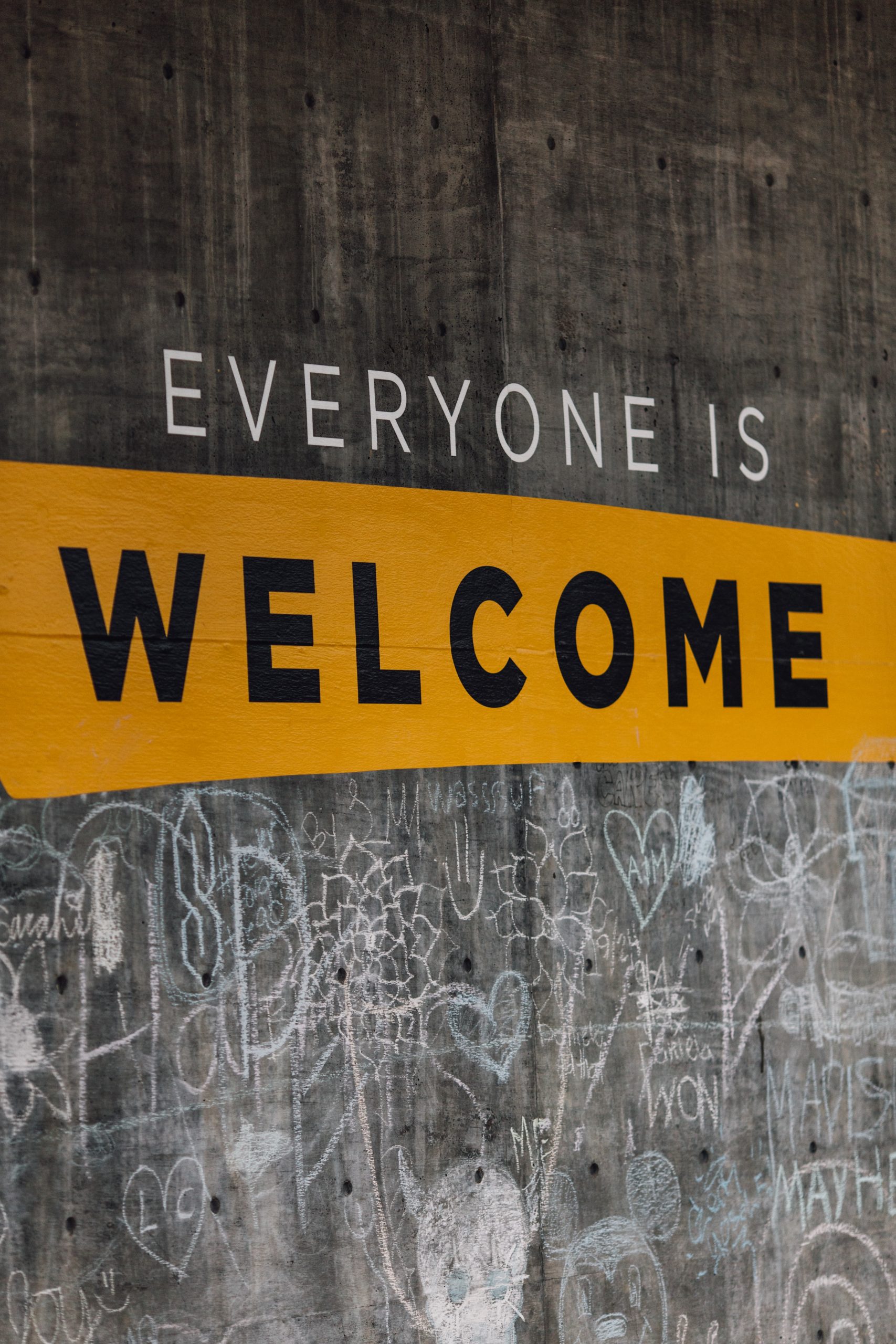 A sign reads everyone is welcome on a grey wall. The word welcome is in black lettering with a yellow stripe hrough it 