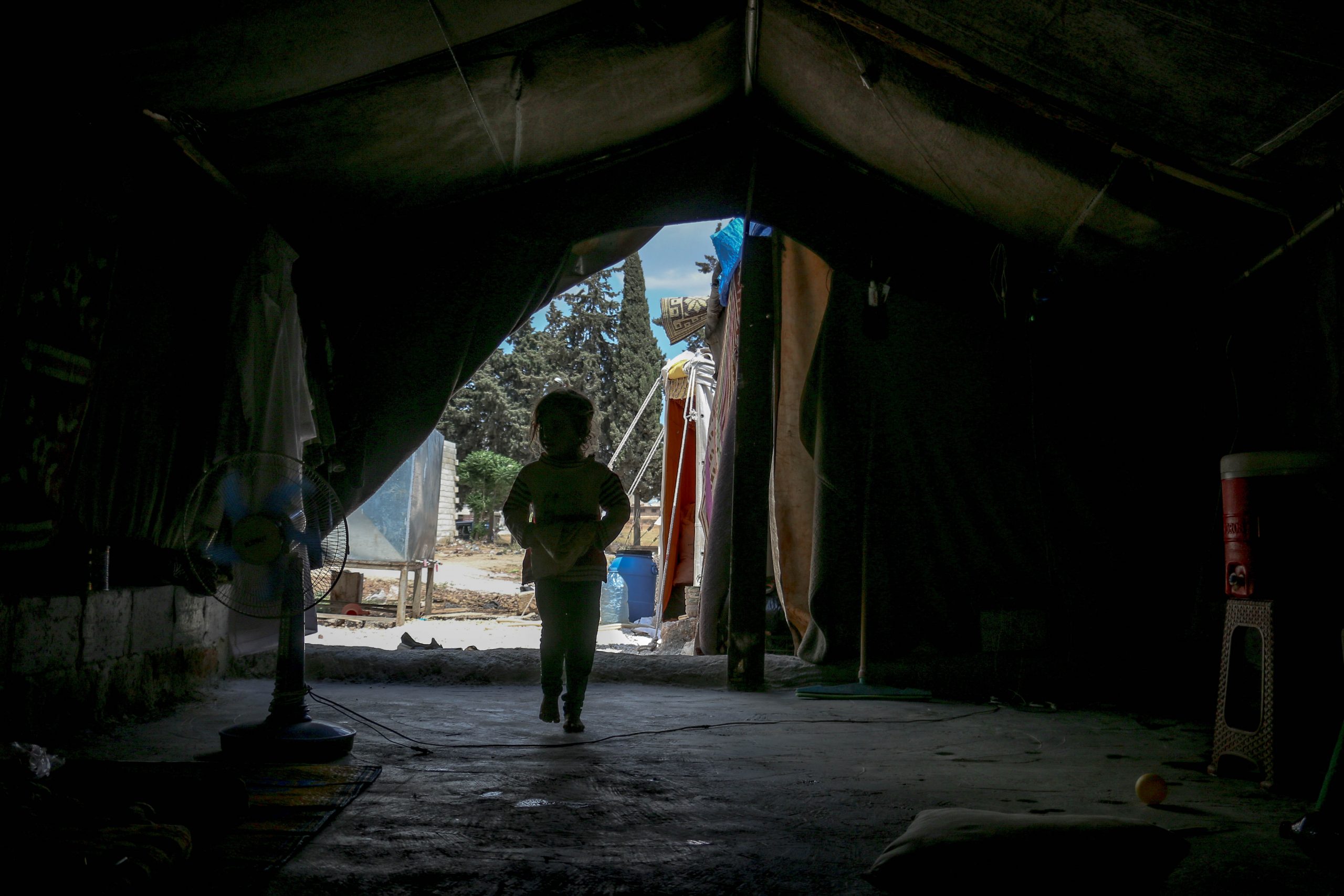 A child refugee is silhouetted beneath a tent. They walk away from the camera shrouded in darkness 