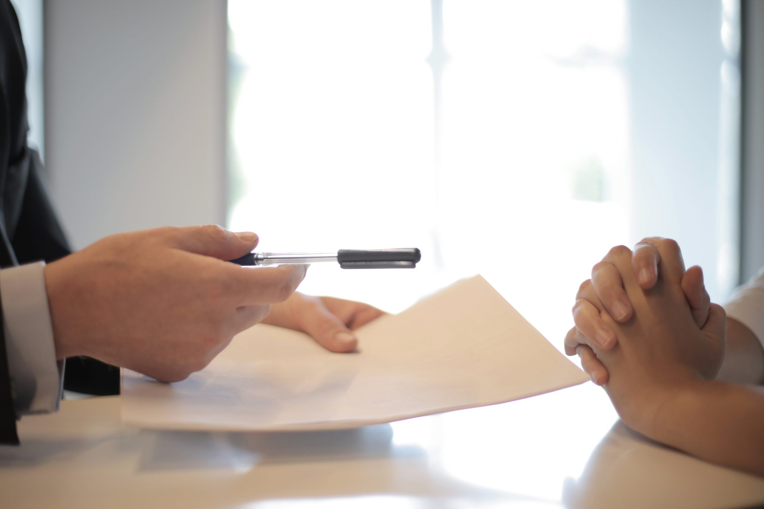 A close up shows a man's hands as he hands a contract for a woman clasping her hands to sign