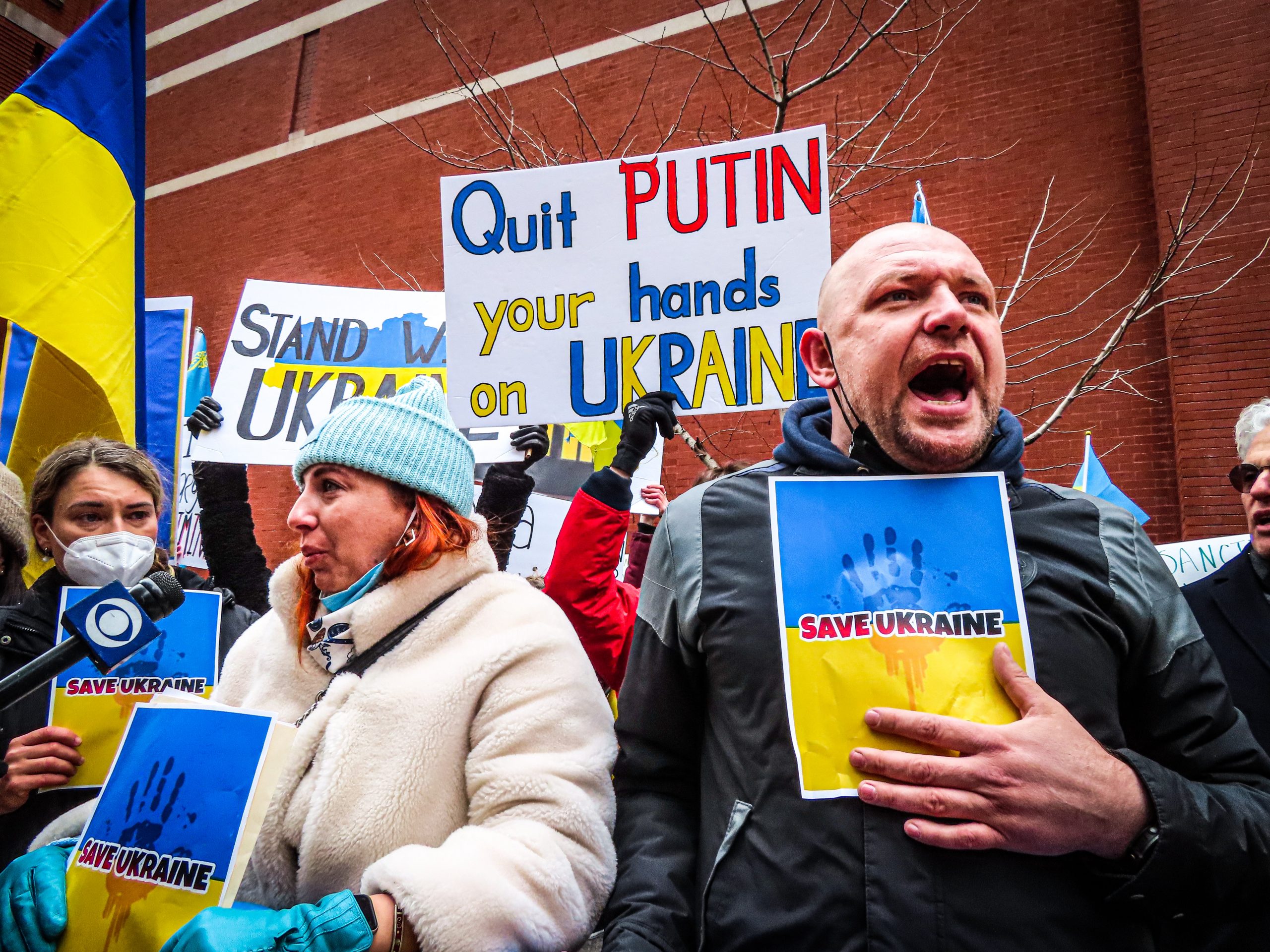 A man shouts during a demonstration for supporting Ukraine. Protesters hold signs denouncing Putin and Ukrainian flags in blue and yellow.