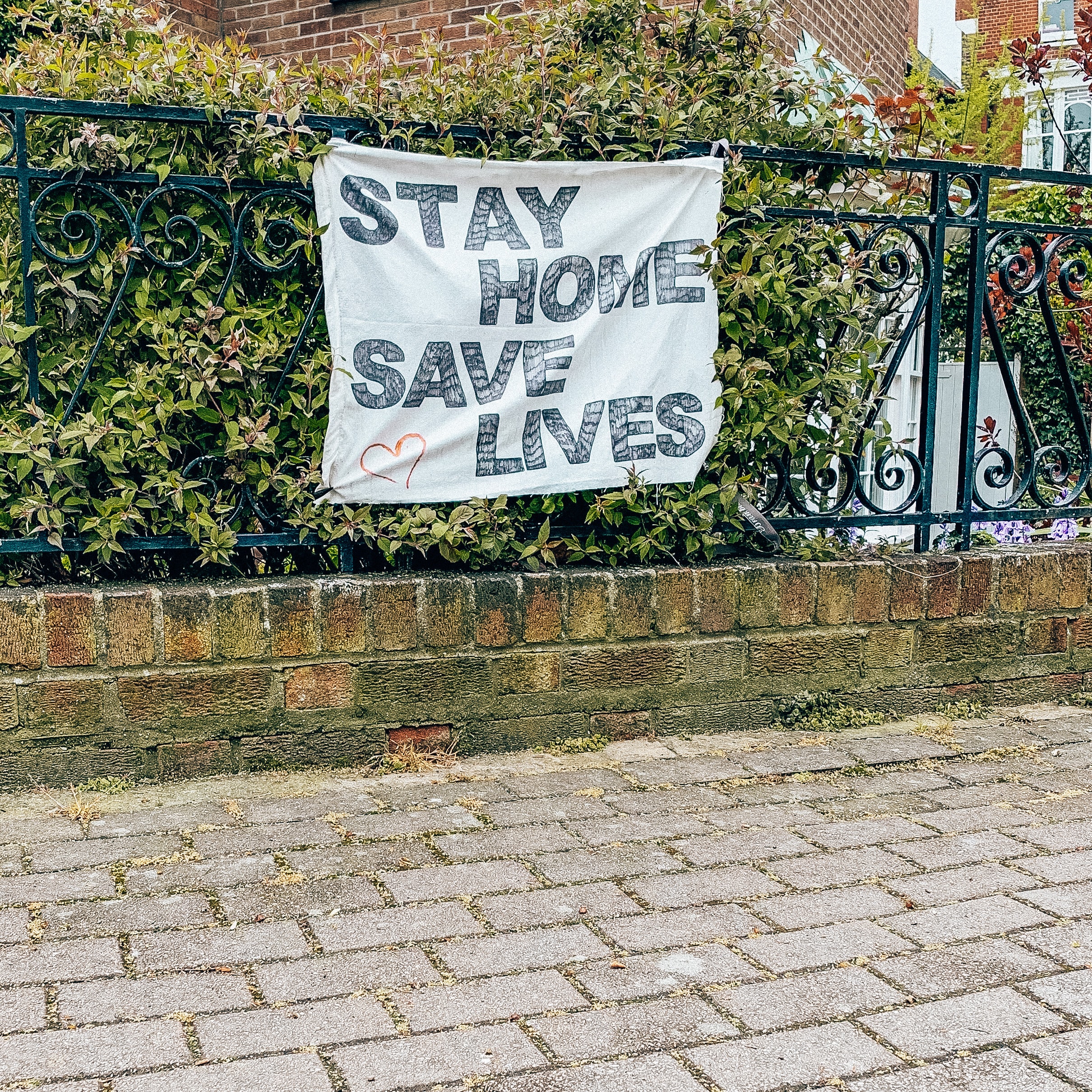 A sign attached to black railing in front of some green shrubbery reads "stay home, save lives" in capital letters in black lettering against a white background. 