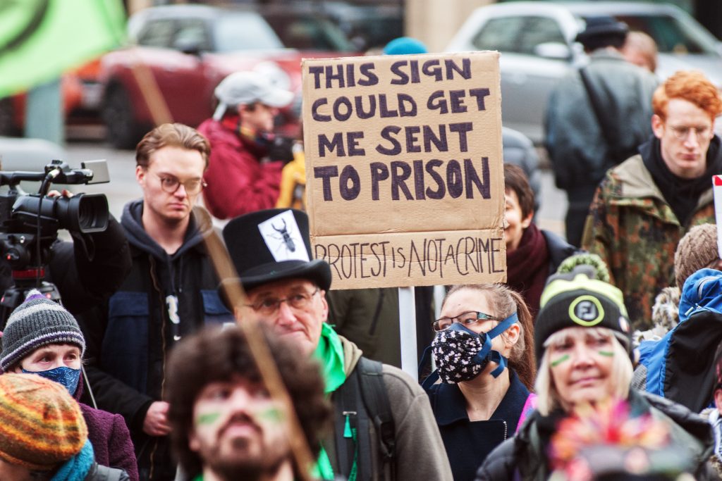 A sign can be seen in a crowd. It reads: "This sign could get me sent to prison. Protest is not a crime."