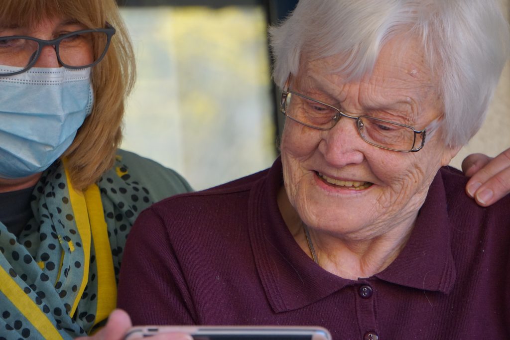 An elderly woman is watching something on a phone being held by a care assistant