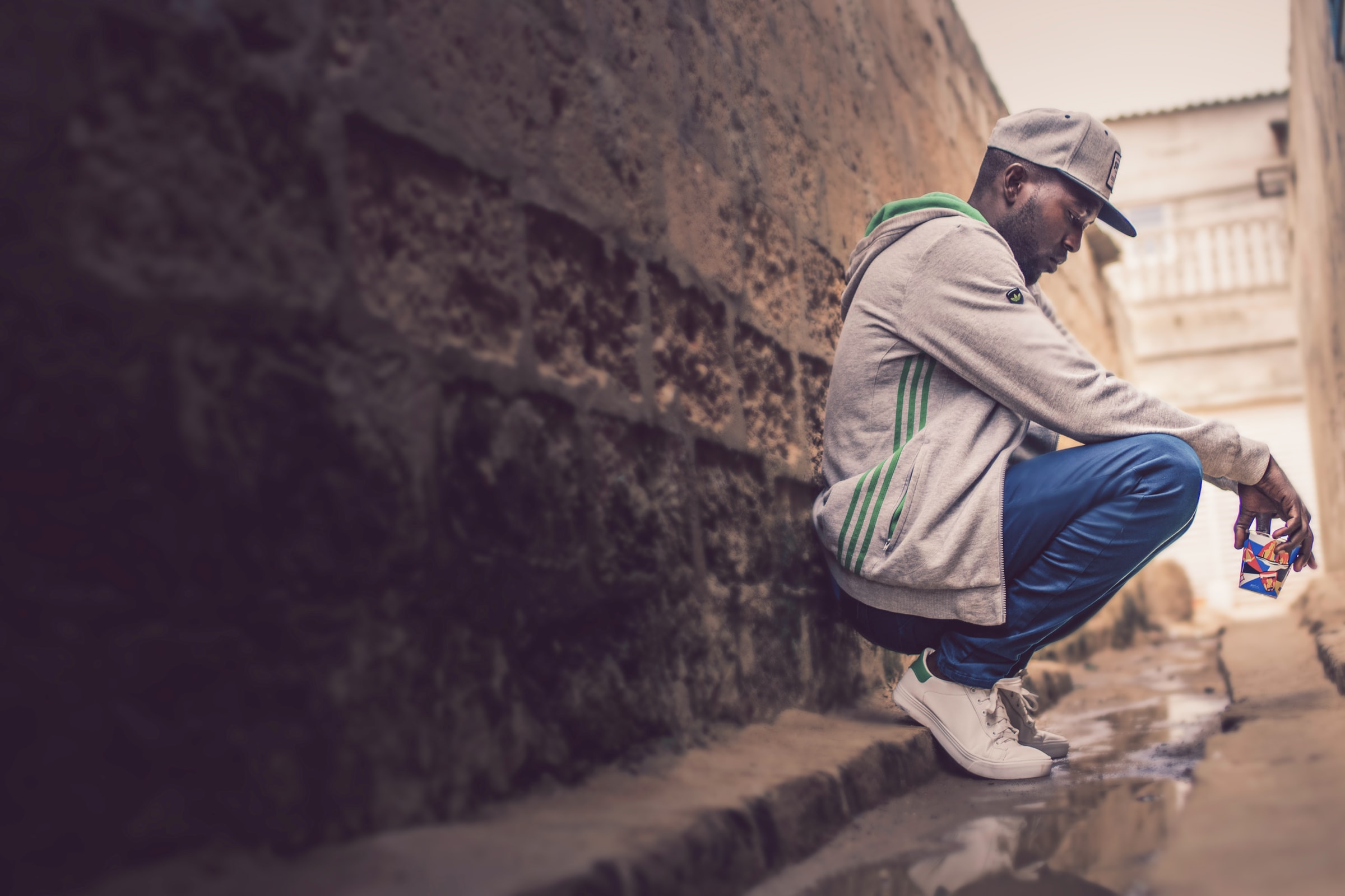A black man wearing a tracksuit and cap squats down in an alleyway, appearing pensive