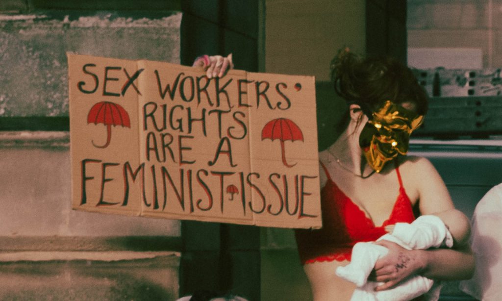 A woman pretends to breastfeed and holds a sign: 'sex workers rights are a feminist issue'