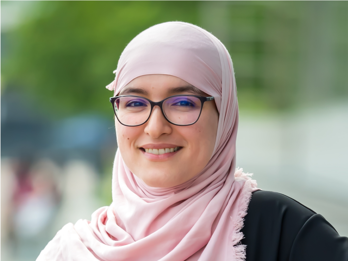 A smiling woman looks at the camera, wearing a pink hijab, a black top and glasses.