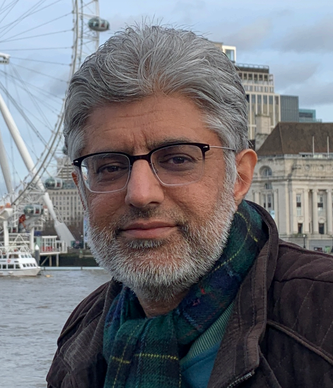 A man with grey hair and a beard, wearing glasses, stands in front of the River Thames looking at the camera.