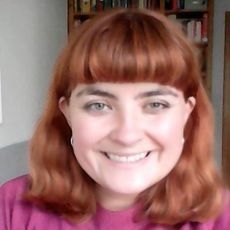 A smiling woman with ginger hair looks into the camera, in a white and grey room with a set of bookshelves, full of books, behind her.