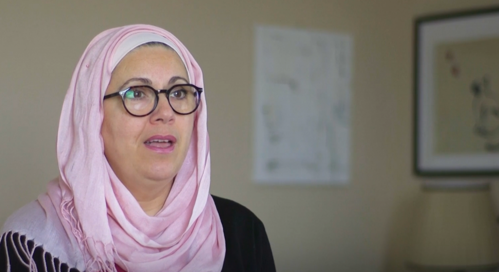 A woman with a pink headscarf, in glasses with black frames is speaking, in an interior room. In the background there are pictures on a light-coloured wall.